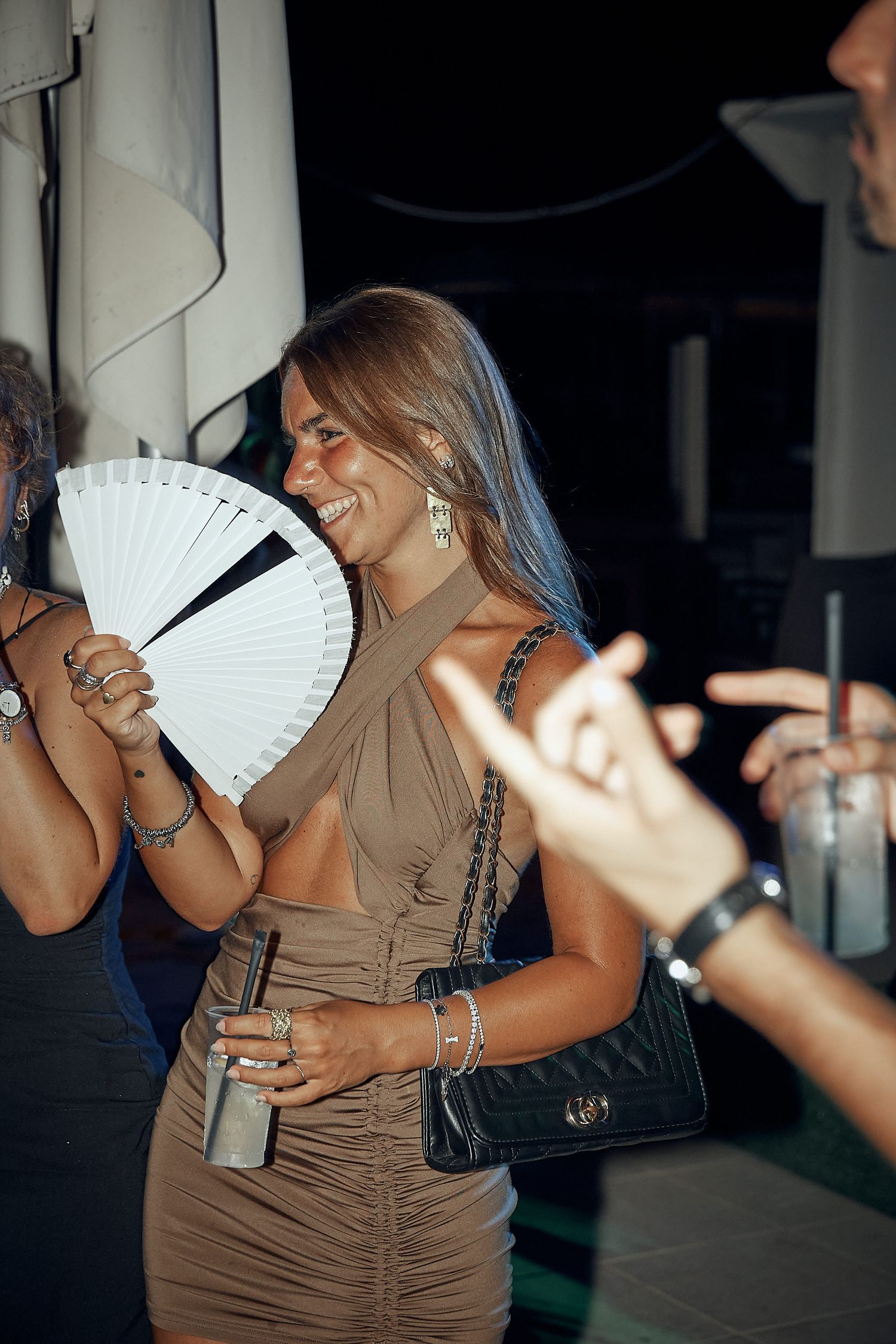 Woman in brown dress holds white fan while dancing at nightclub.