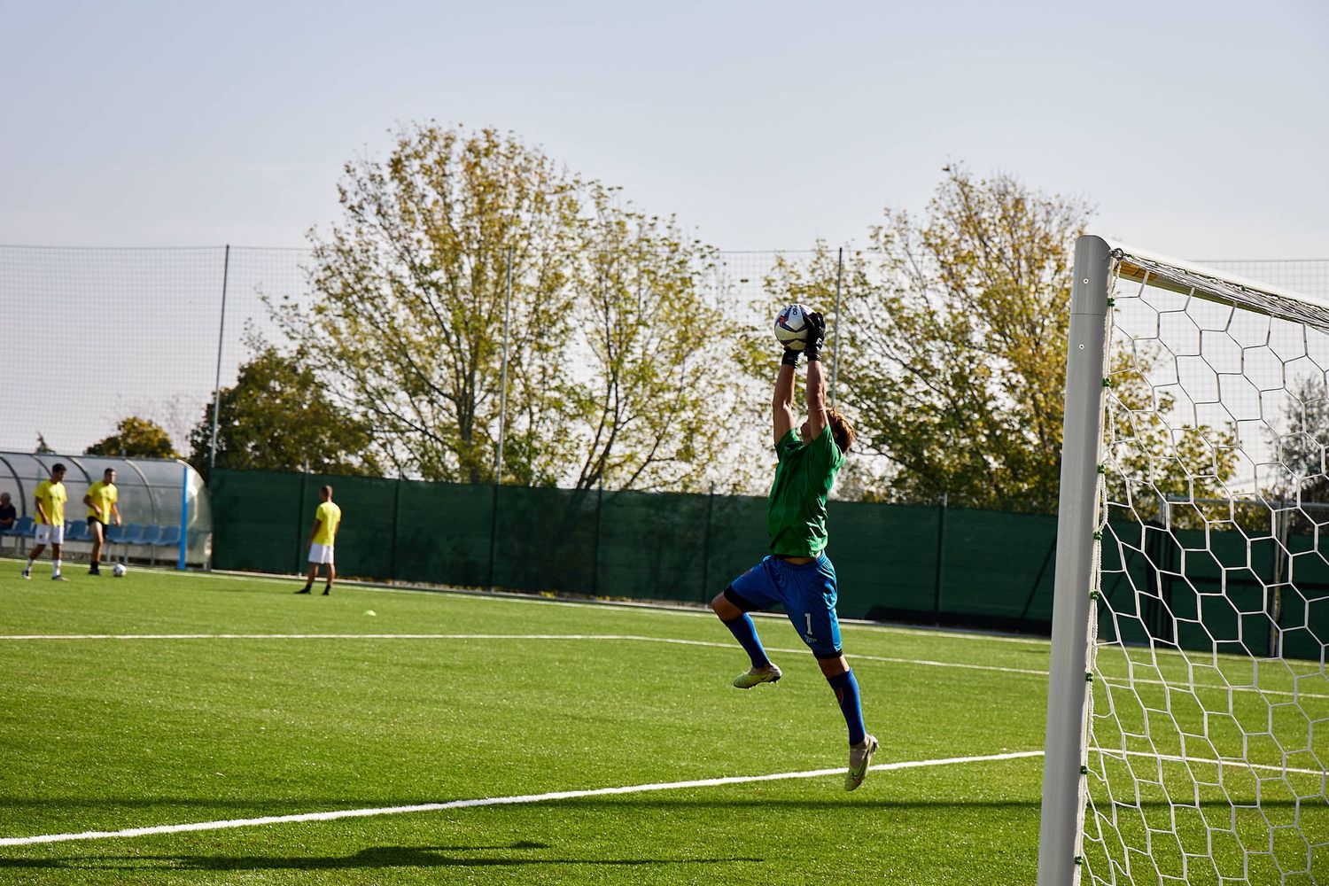 Soccer goalkeeper jumps high to defend goal post during match on sunny day.