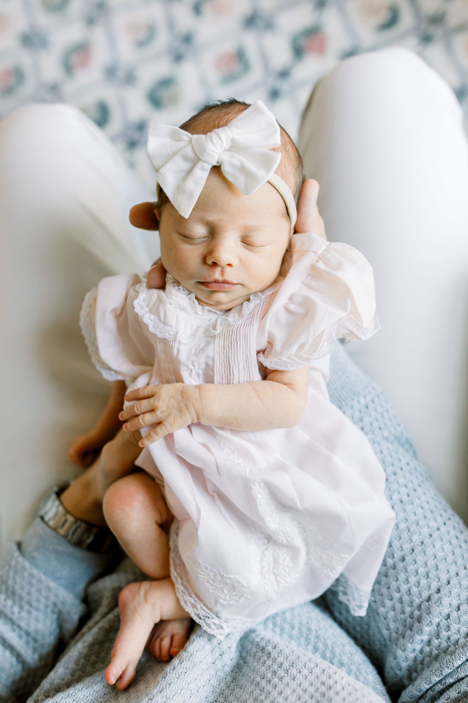 Sleeping newborn baby wearing white dress and bow headband on blue blanket.