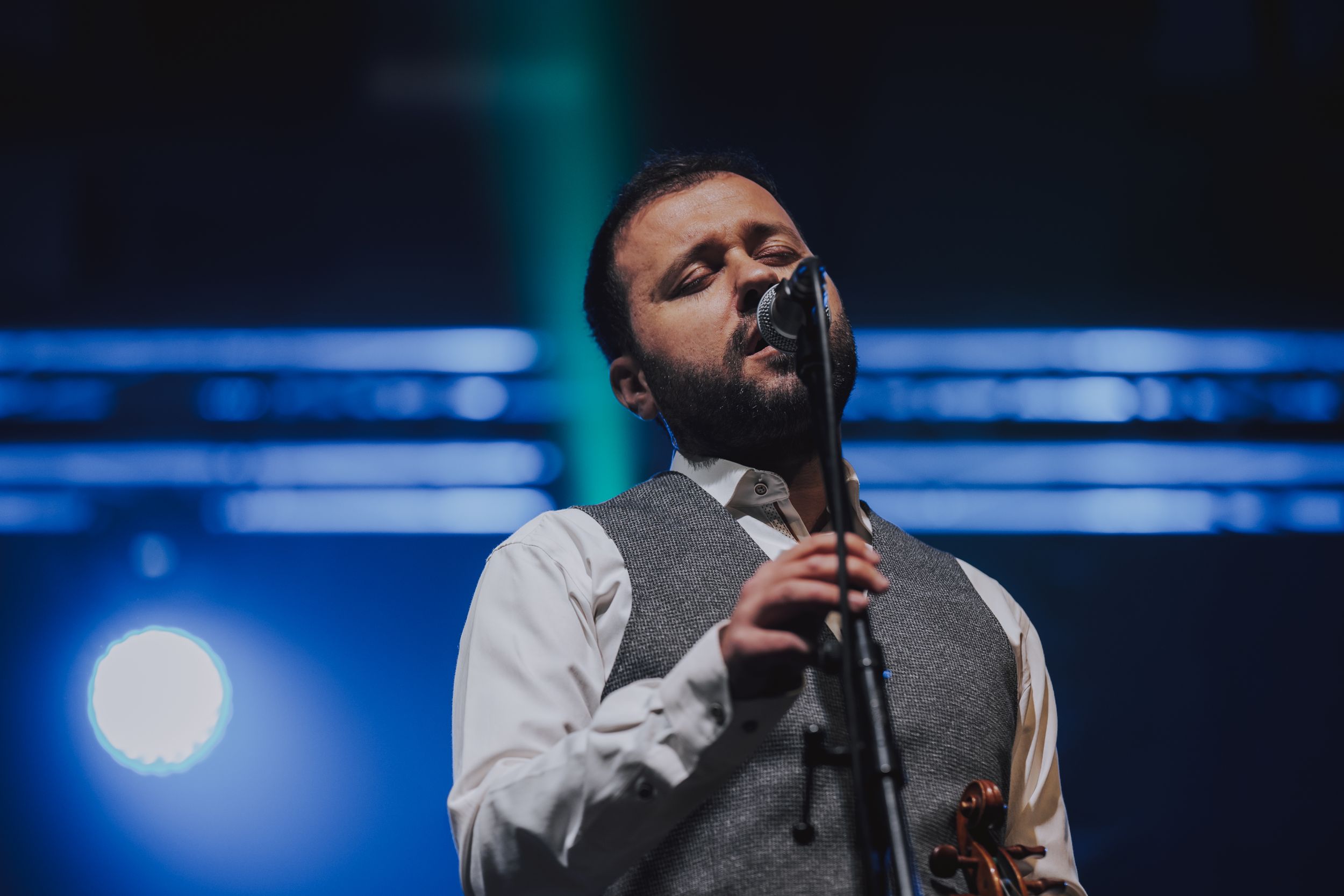 Performer in grey vest singing emotionally into microphone under blue stage lighting.