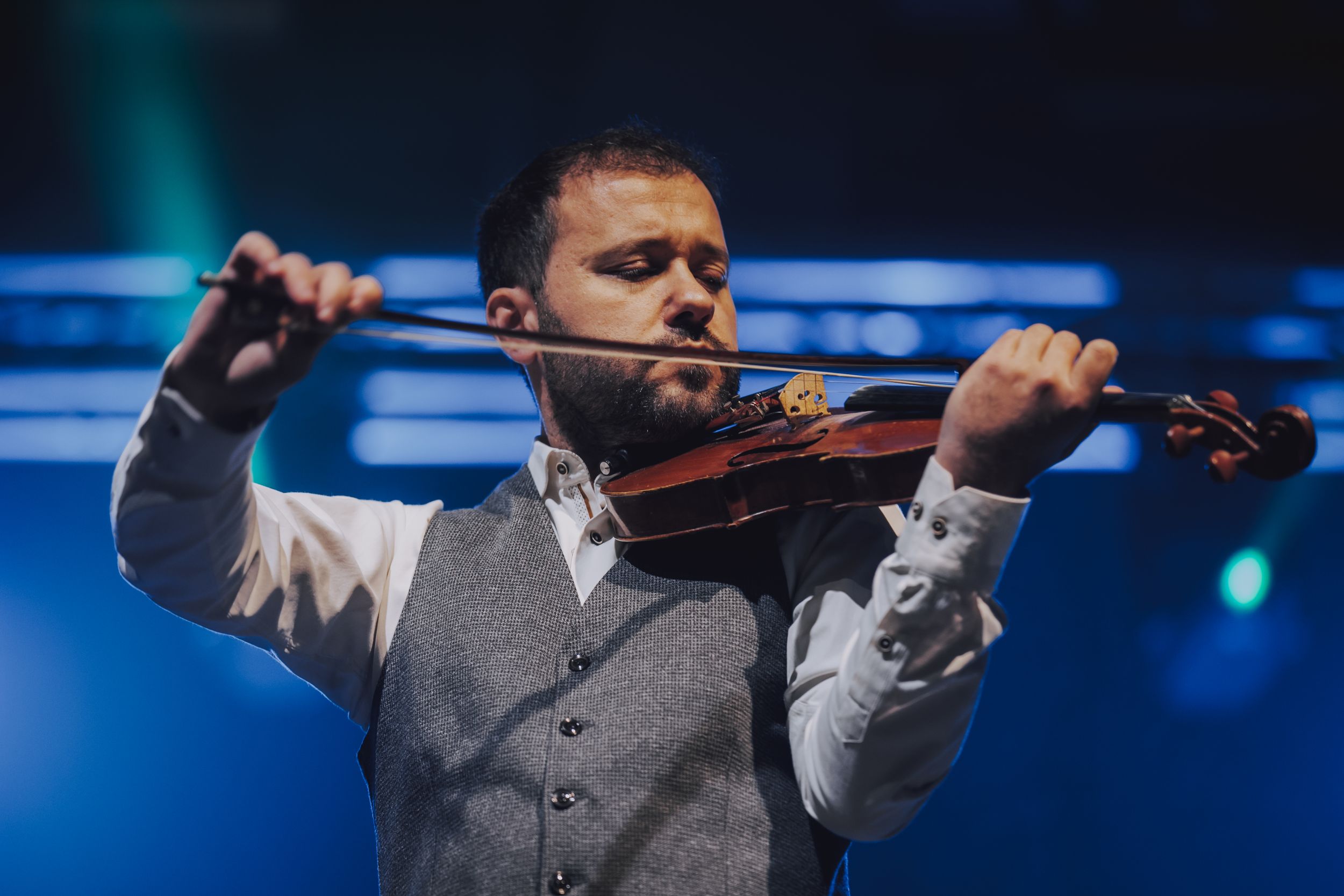 Musician in vest and white shirt playing violin during stage performance.