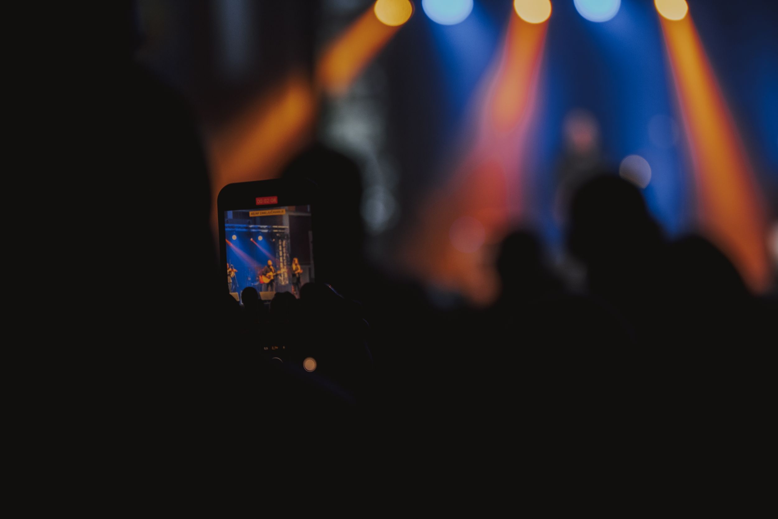 Silhouetted phone screen capturing concert performance with colorful stage lights in background.