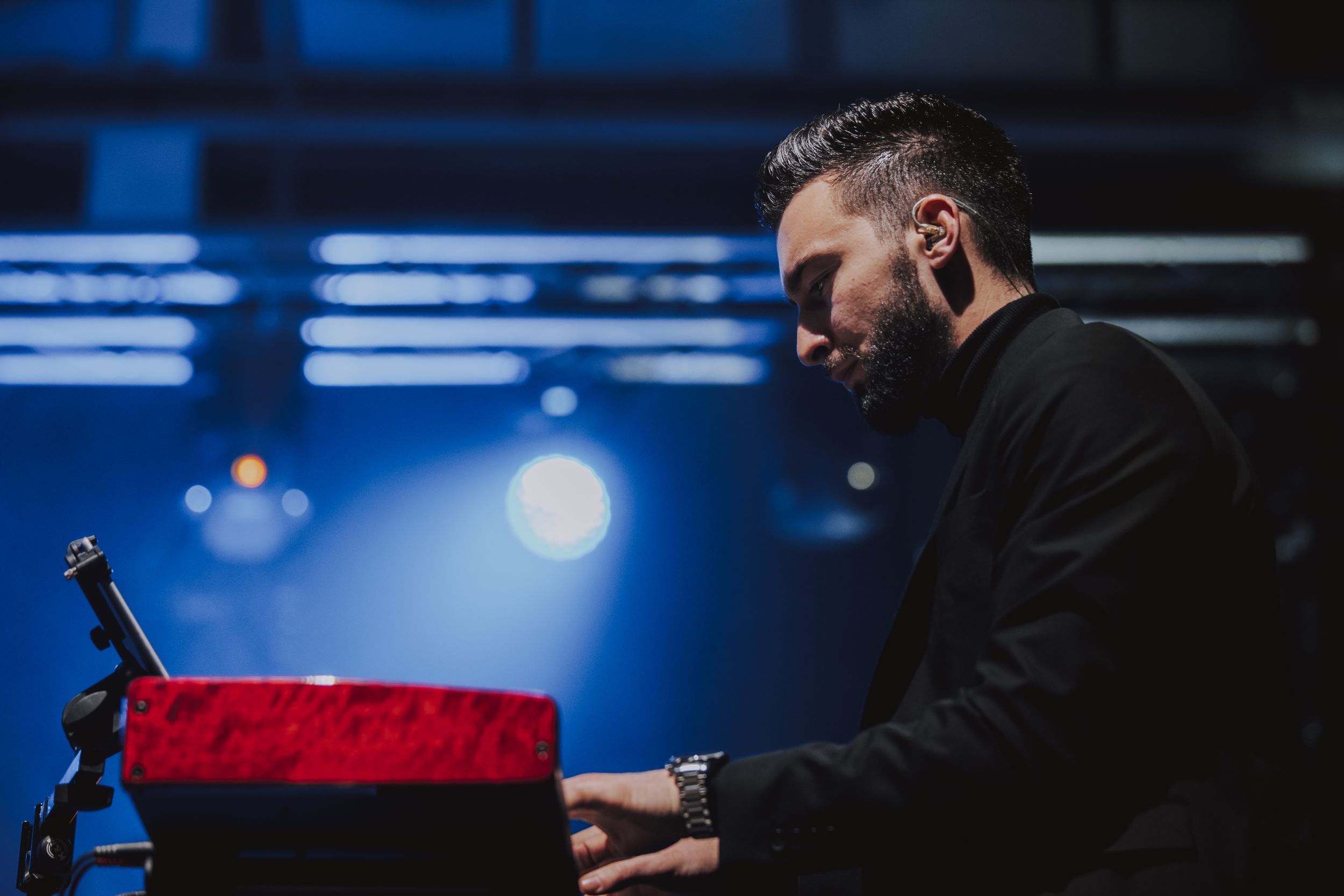 Musician performs at red keyboard during concert with blue stage lighting.