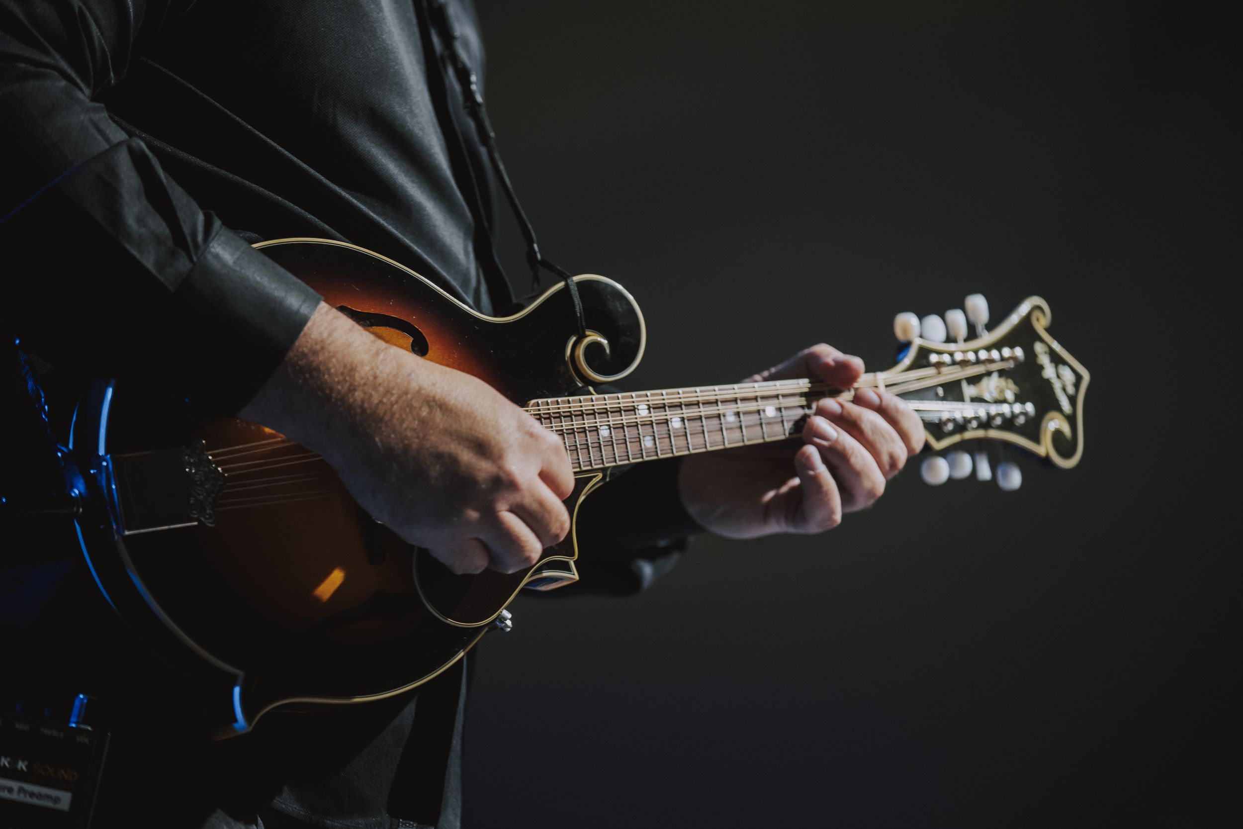 Close-up of hands playing mandolin strings against dark background.