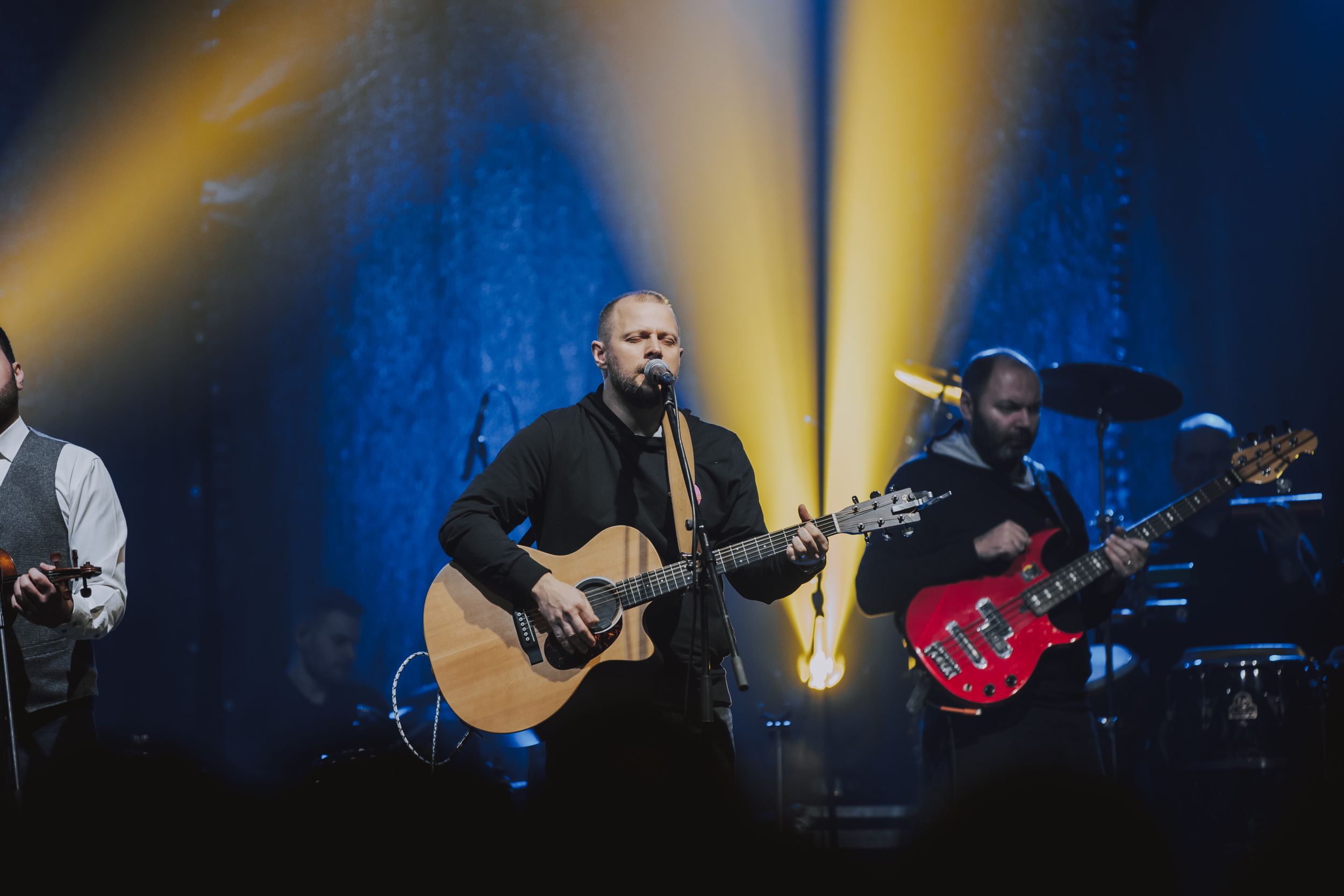 Band performs on concert stage with acoustic guitar and dramatic yellow lighting effects.