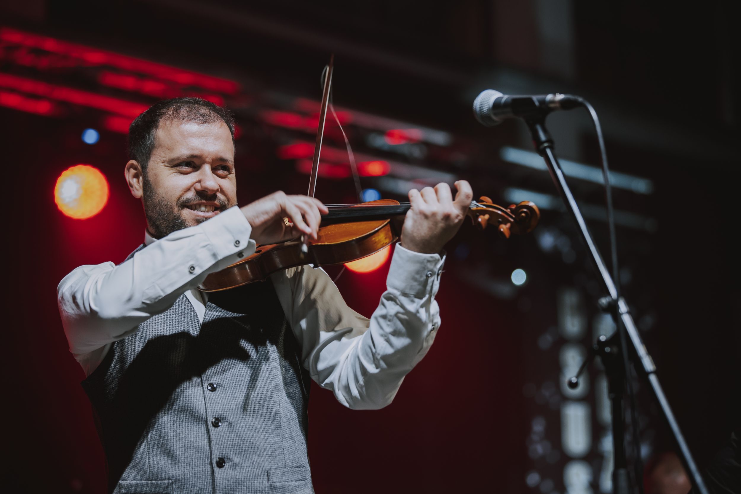 A musician playing violin on stage wearing a white shirt and grey vest under dramatic red lighting.