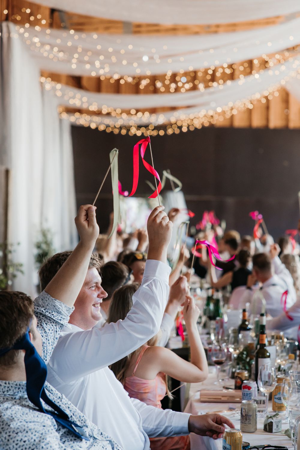 Wedding reception guests raise hands in celebration under twinkling string lights.