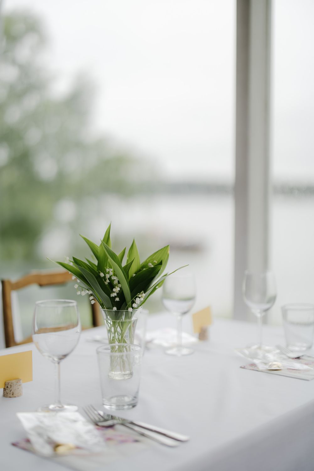 Elegant wedding table setting with lily of the valley centerpiece, wine glasses, and silverware on white linen near window.