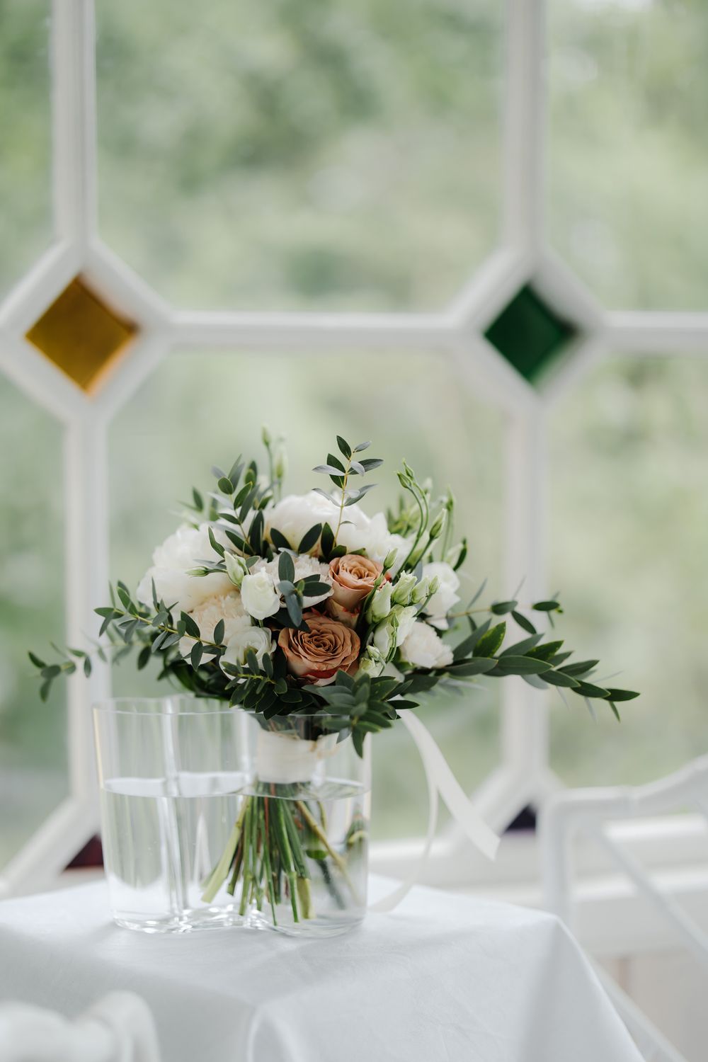Elegant wedding bouquet of white and blush roses with greenery in a glass vase by a stained glass window.