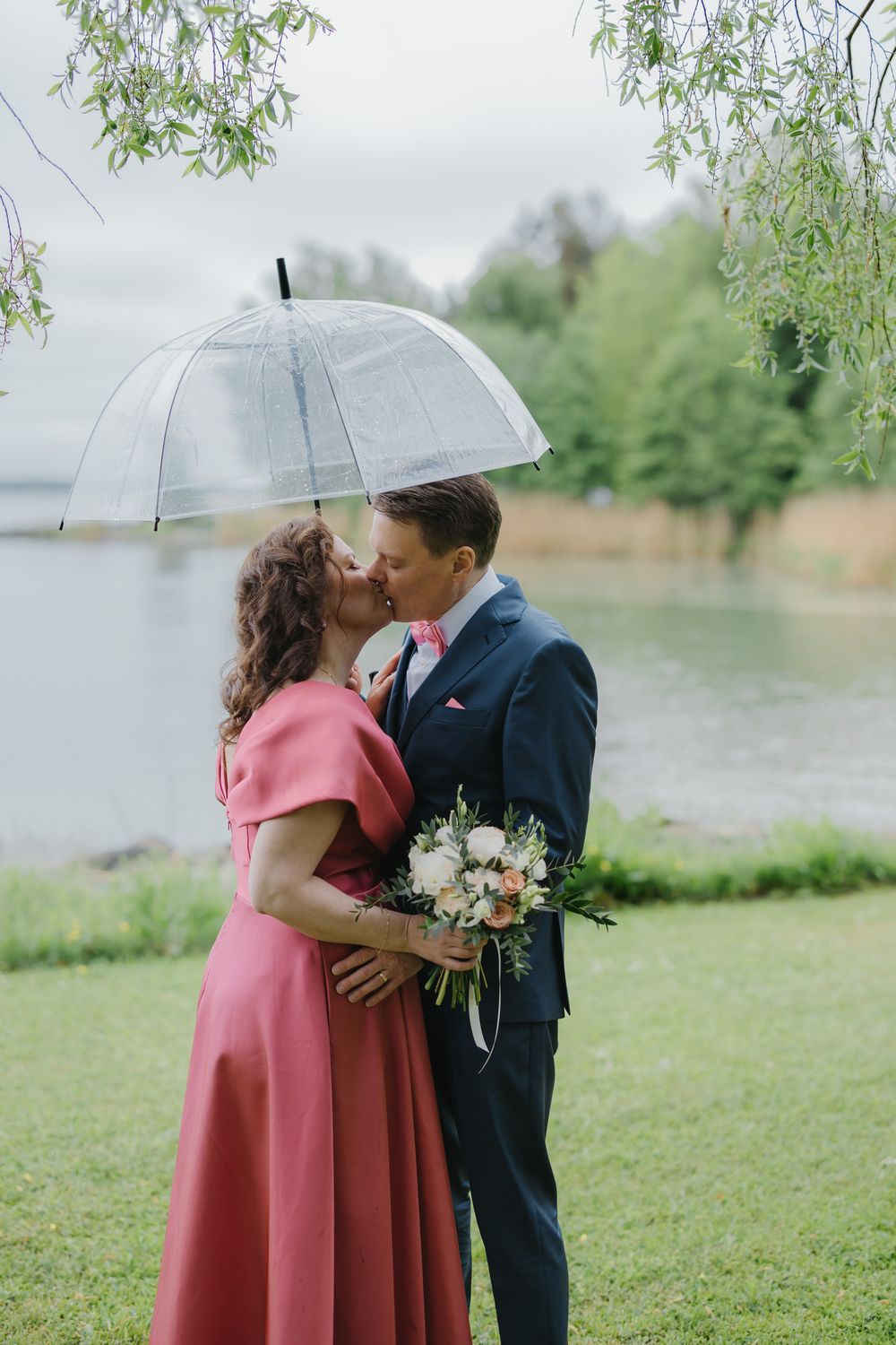 Couple kissing under a clear umbrella in the rain, bride in pink dress holding bouquet, groom in navy suit by a lake.
