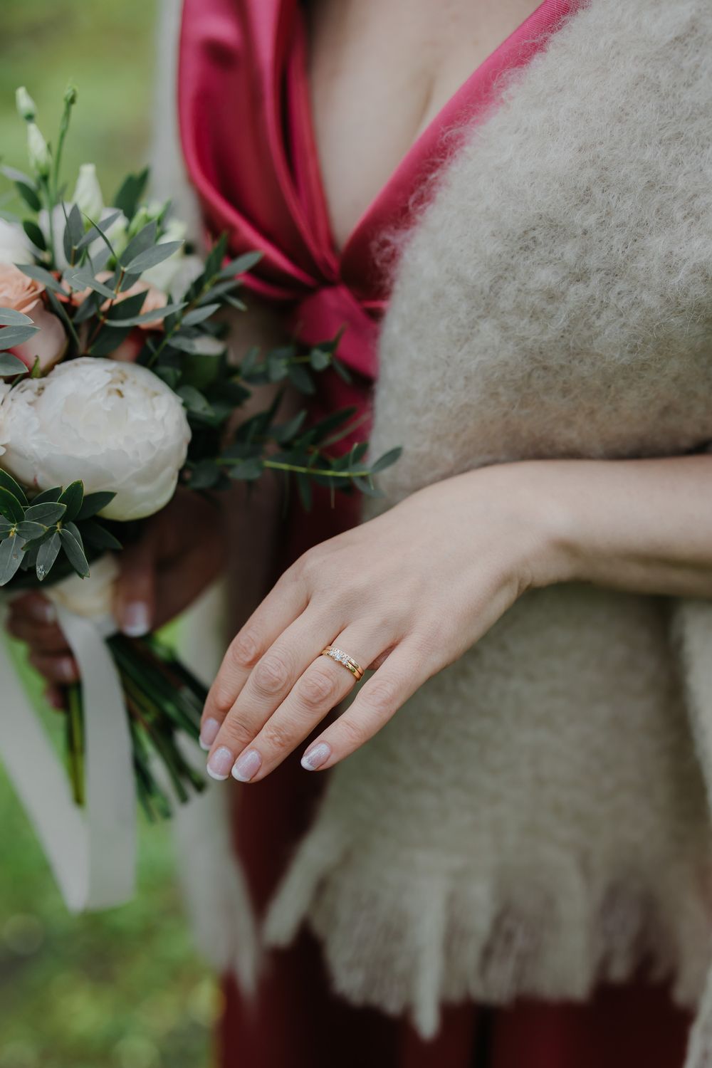 Close-up of a bride's hand wearing a gold wedding band, holding a white peony bouquet with greenery.