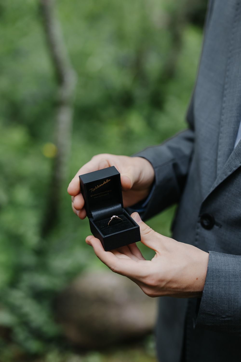 Man in gray suit holding open black ring box with diamond engagement ring outdoors in green forest setting.