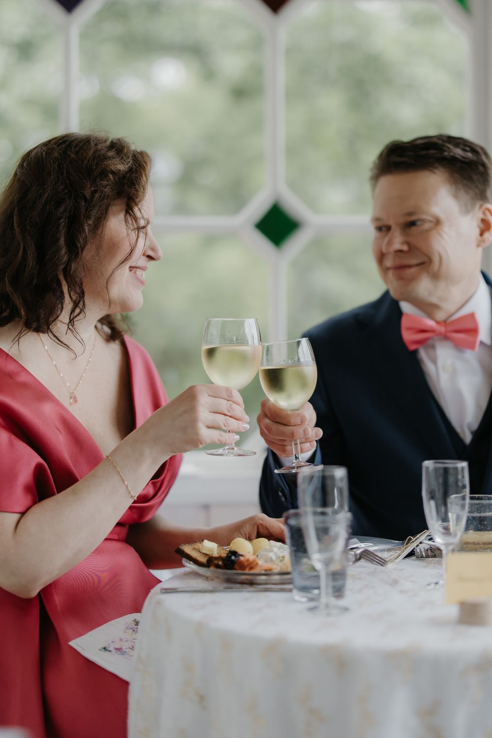 Couple in formal attire toasting with white wine glasses at a wedding reception table near stained glass windows.