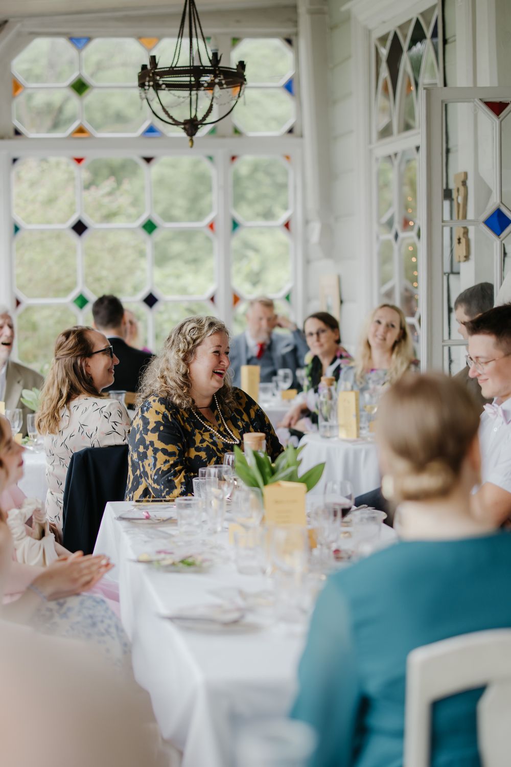 Guests laughing and celebrating at a wedding reception in an elegant venue with colorful stained glass windows.