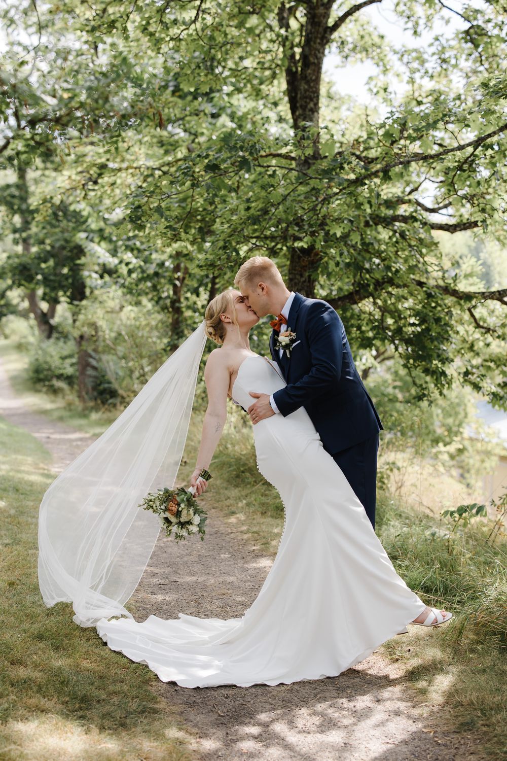 Bride and groom share a romantic dip kiss outdoors, with flowing veil and bridal bouquet, surrounded by lush green trees.