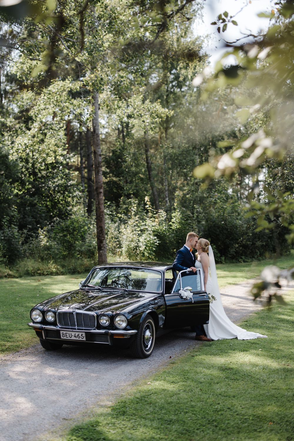 Bride and groom kissing beside a classic black Jaguar car surrounded by lush green trees on their wedding day.