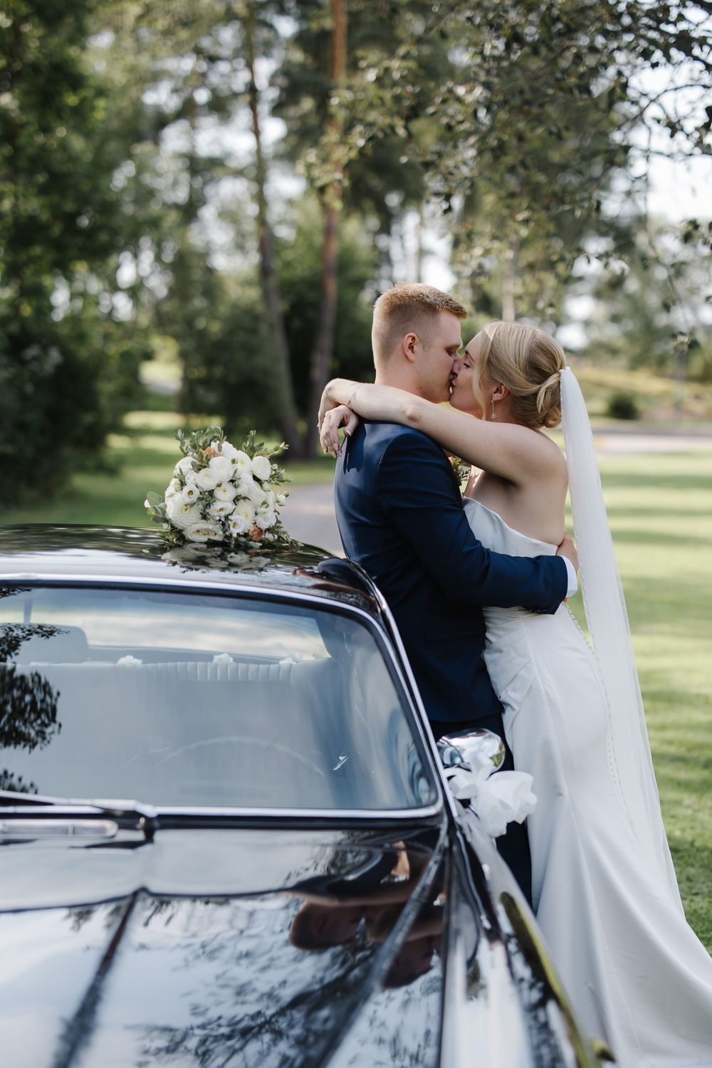Bride and groom kissing beside a vintage black car, holding a white bouquet, surrounded by lush green trees.
