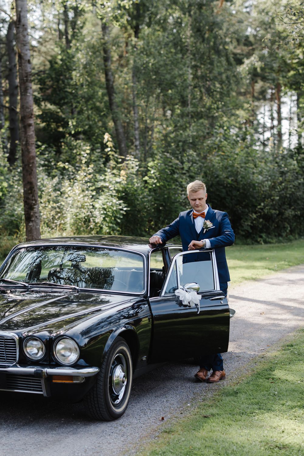 Groom in navy suit with orange bow tie stands beside a classic black Jaguar car on a tree-lined driveway.