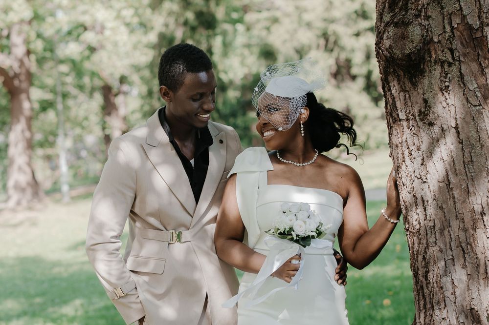 A joyful couple in wedding attire smiles at each other outdoors, with the bride in white and groom in a beige suit.
