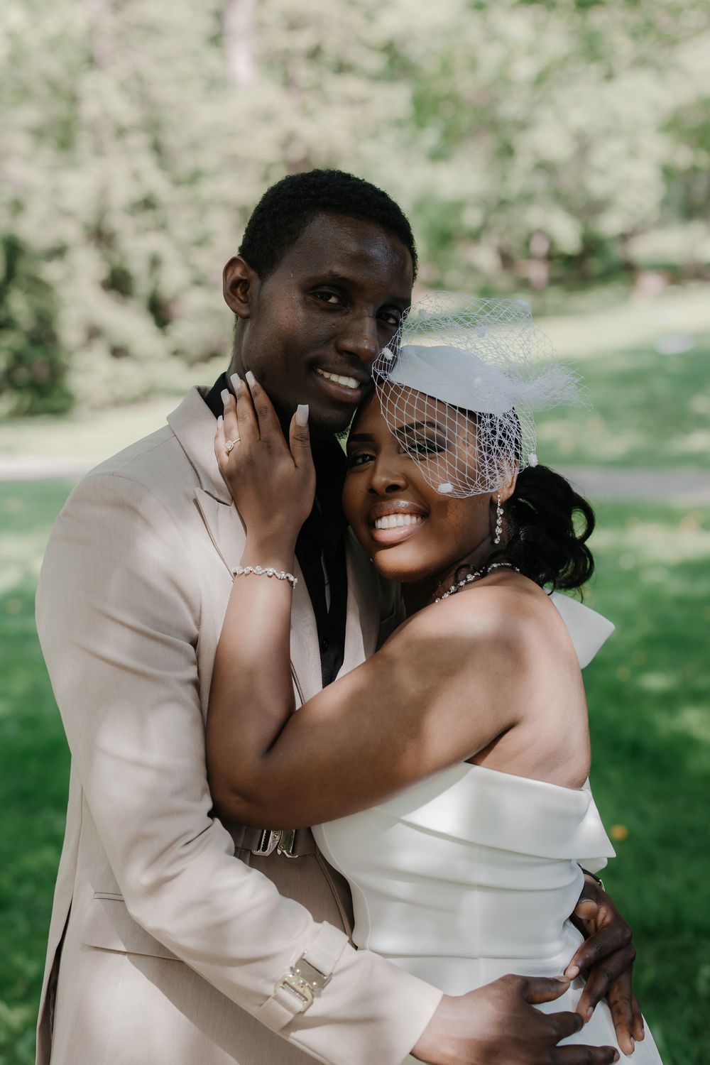 A joyful Black couple in wedding attire embraces outdoors, groom in beige suit, bride in white dress with birdcage veil.