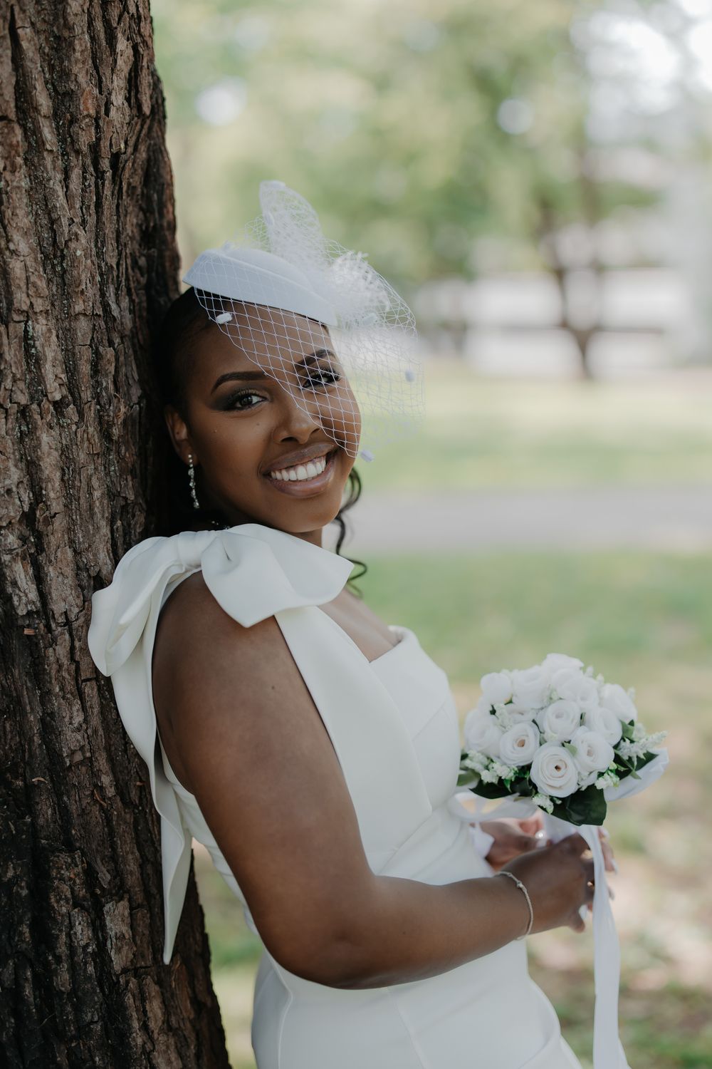 Smiling Black bride in white dress and birdcage veil holds rose bouquet, leaning against tree in park.
