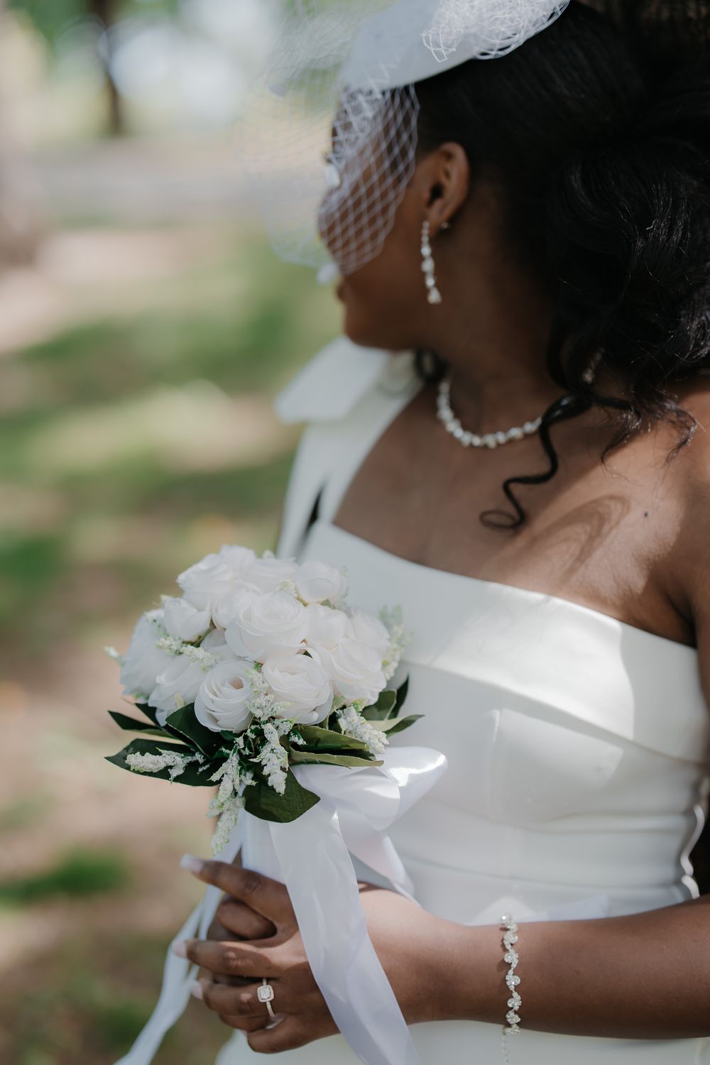 Bride in white off-shoulder gown holding elegant white rose bouquet with ribbon, wearing pearl jewelry and veil.