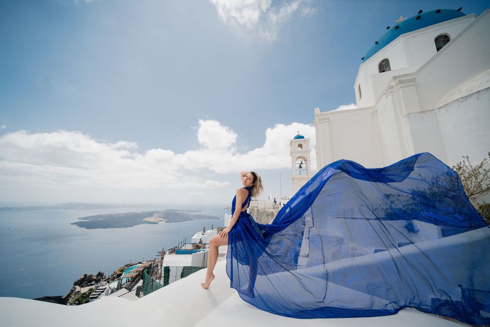 Flowing blue dress creates dramatic silhouette against white architecture and Aegean Sea in Santorini.