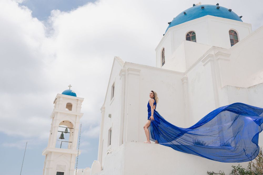 Flowing blue dress catches wind by iconic white church with blue dome in Santorini.