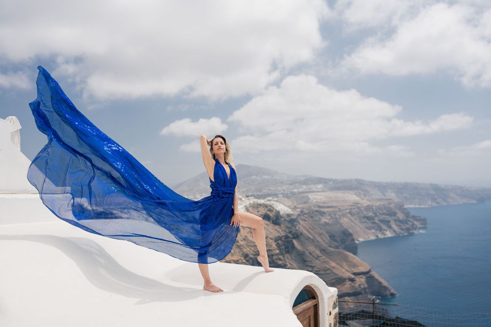 Billowing blue dress catches wind on white rooftop overlooking Mediterranean coastline.