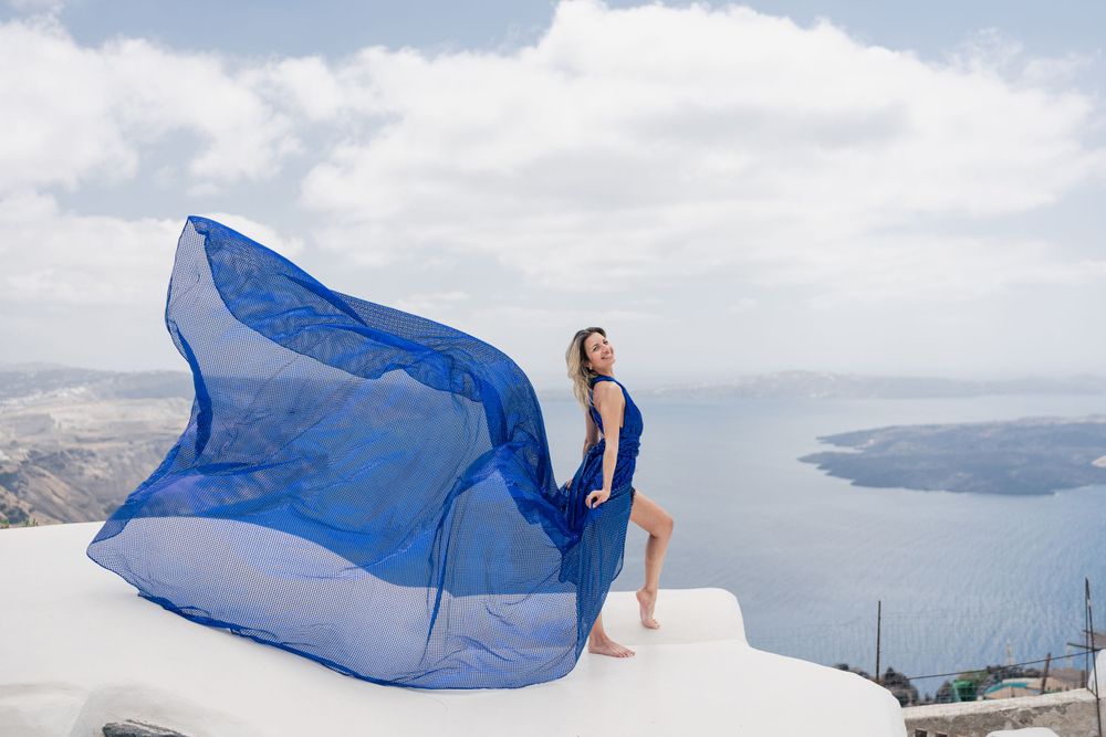Flowing blue fabric catches the wind against a stunning Santorini backdrop with white buildings and sea views.