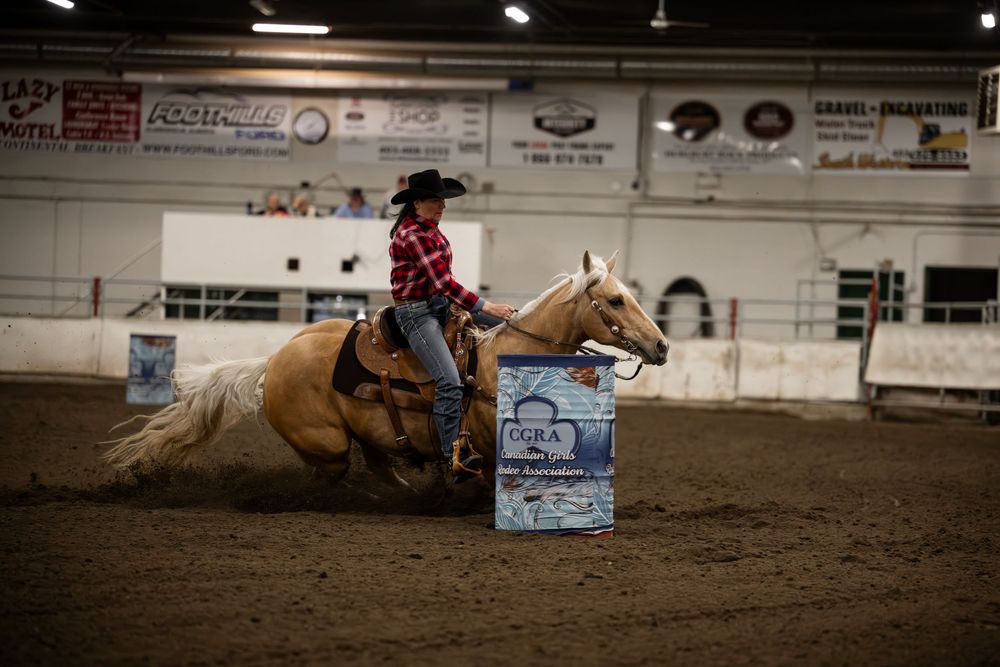 A rodeo rider competes in barrel racing, guiding their horse in a tight turn around a barrel in an indoor arena.