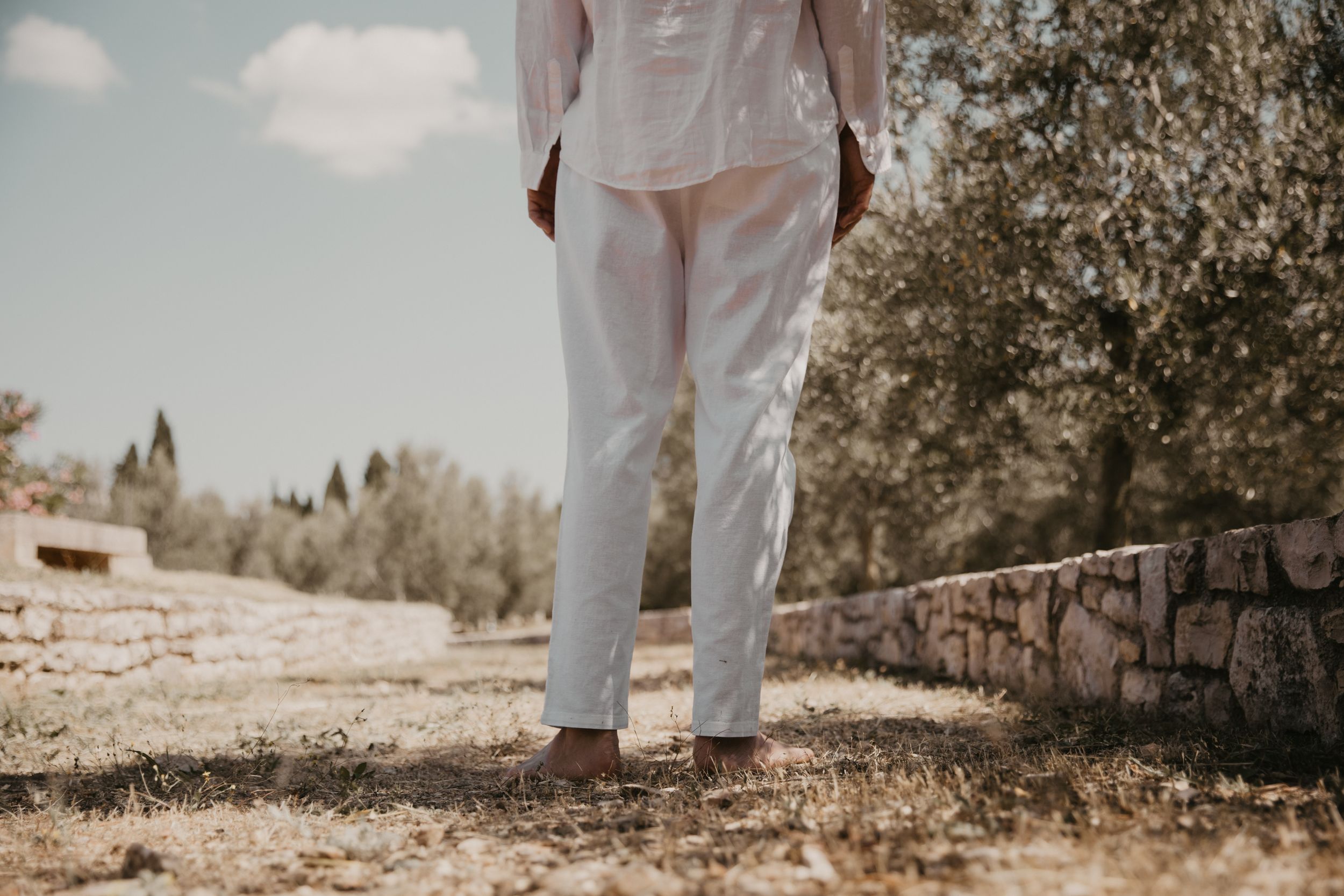 Person in white linen outfit stands near stone wall on sunny path.