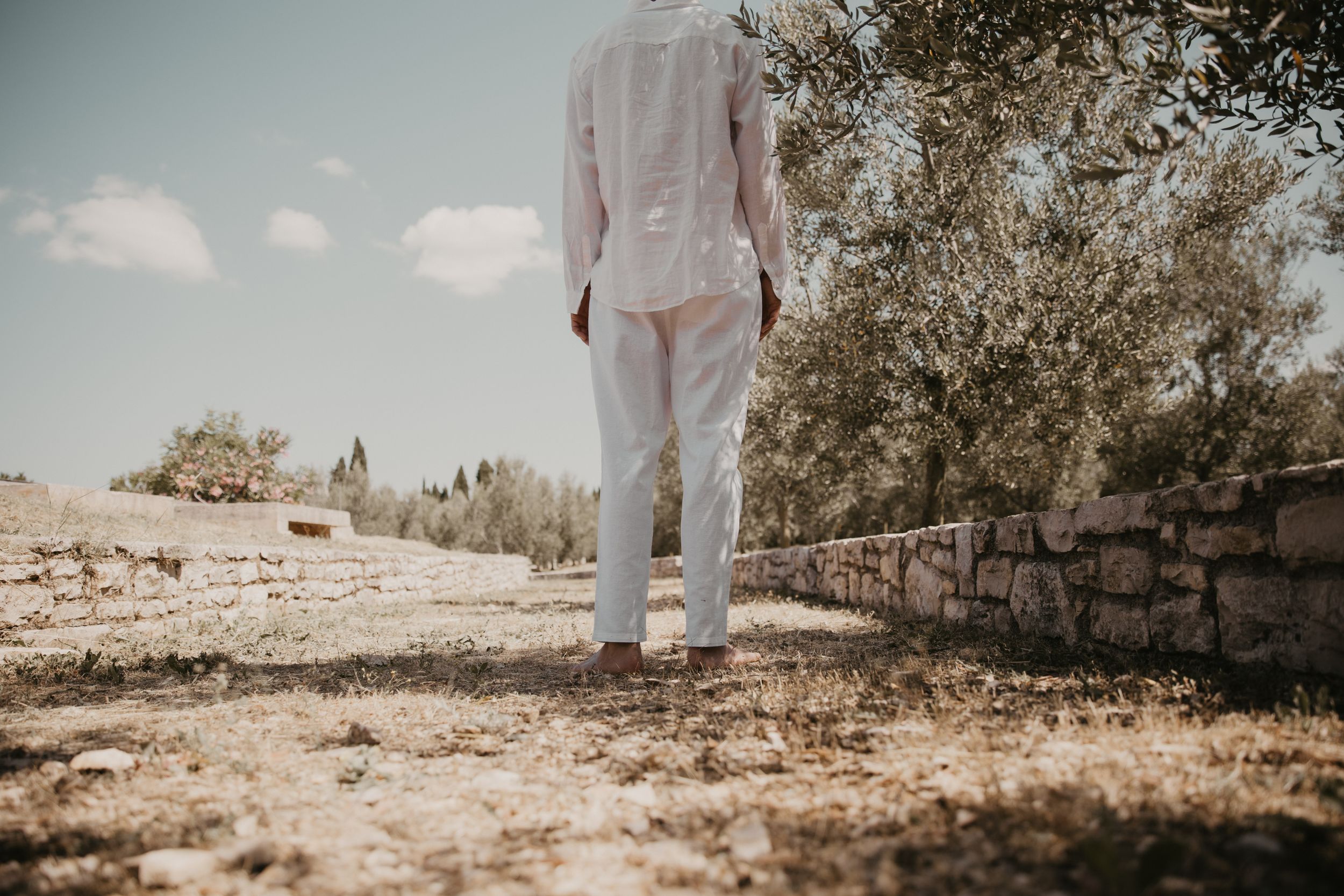 Figure in white clothing stands by rustic stone wall on dusty path.