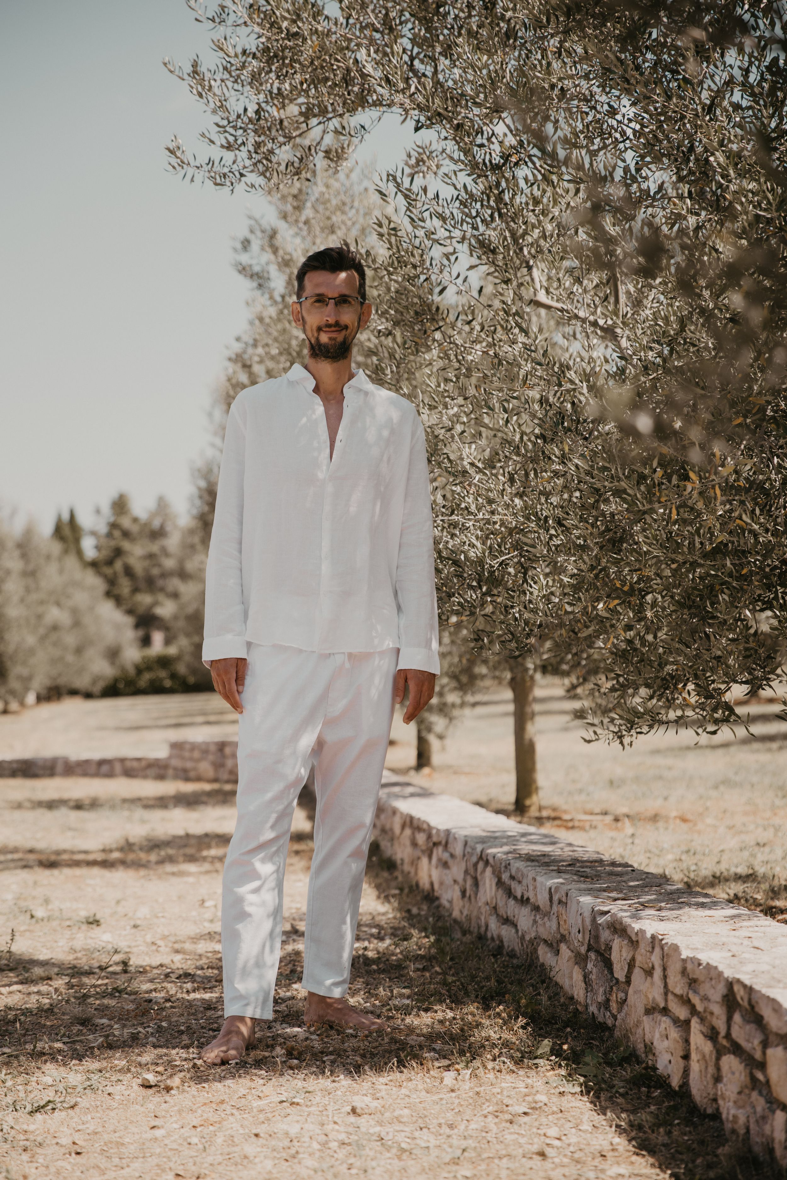 Person in white linen clothing poses on stone wall path surrounded by trees.