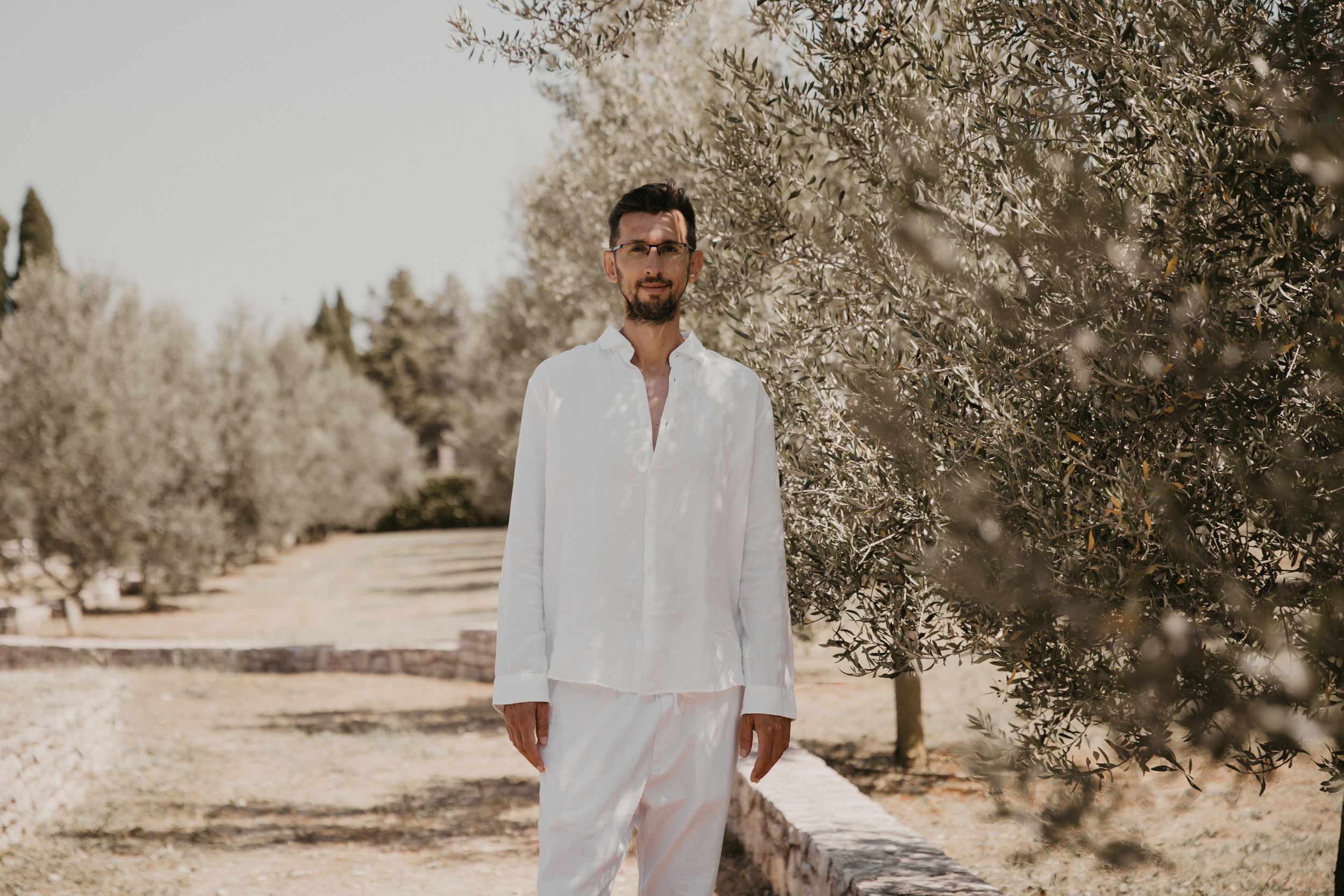 Person in crisp white linen outfit stands on stone path among olive trees.