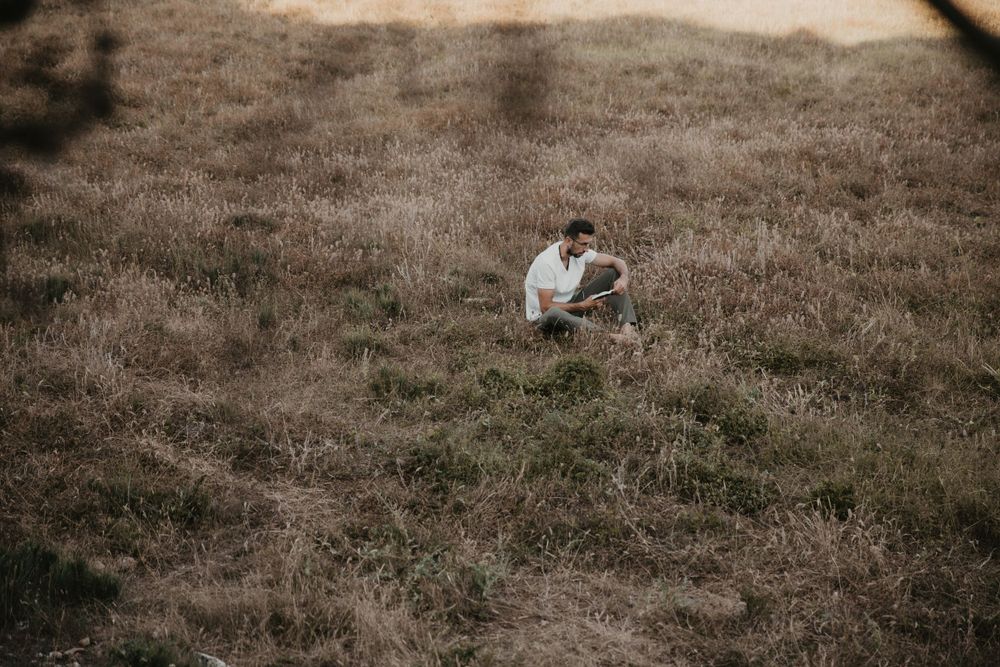 A solitary figure crouches thoughtfully on a grassy hillside at dusk.