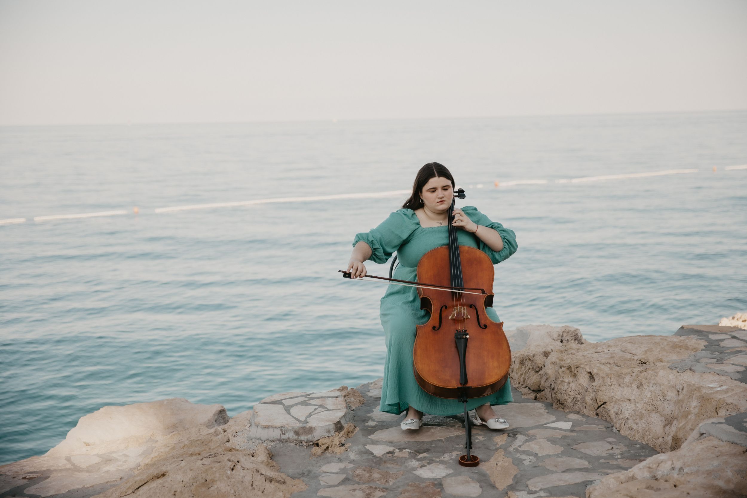 A cellist in a teal dress performs on rocky cliffs overlooking a serene ocean view.