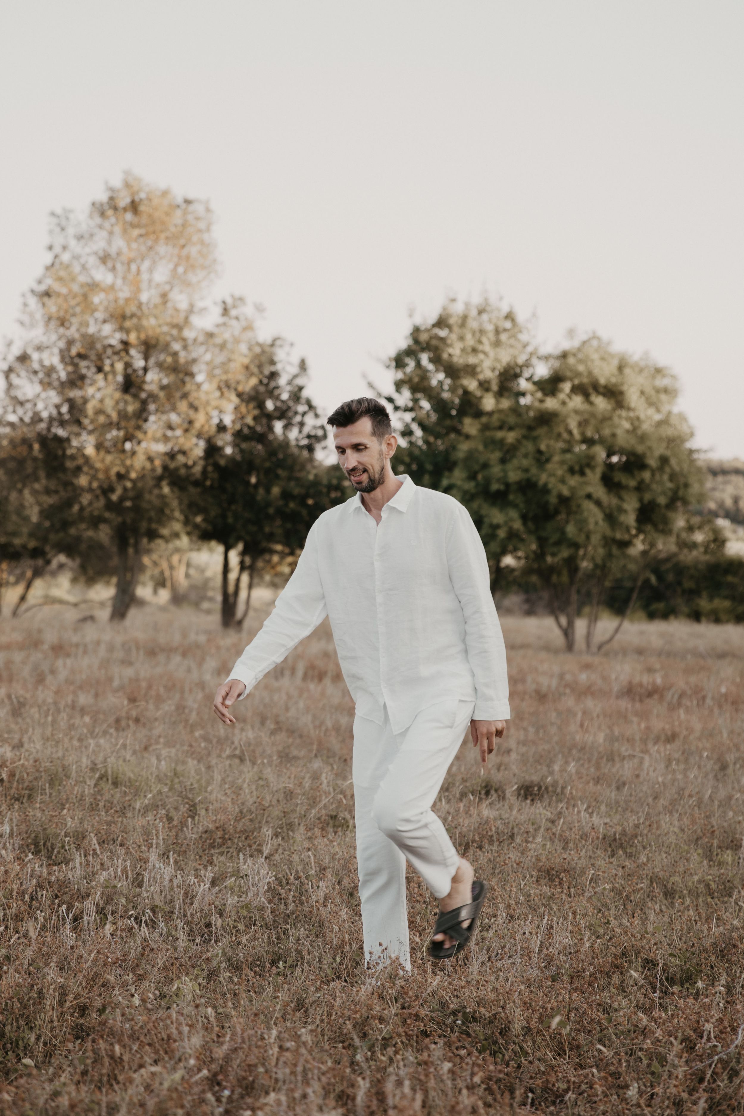 Person in white linen outfit walking through rustic field with trees.