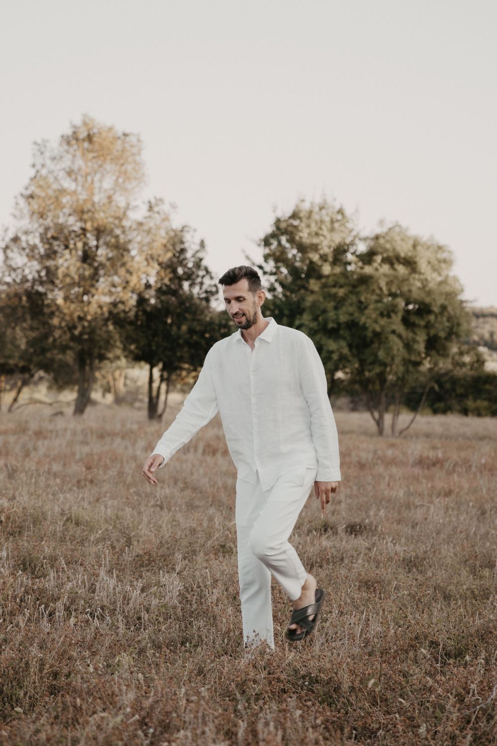 Person in white linen outfit walking through rustic field with trees.
