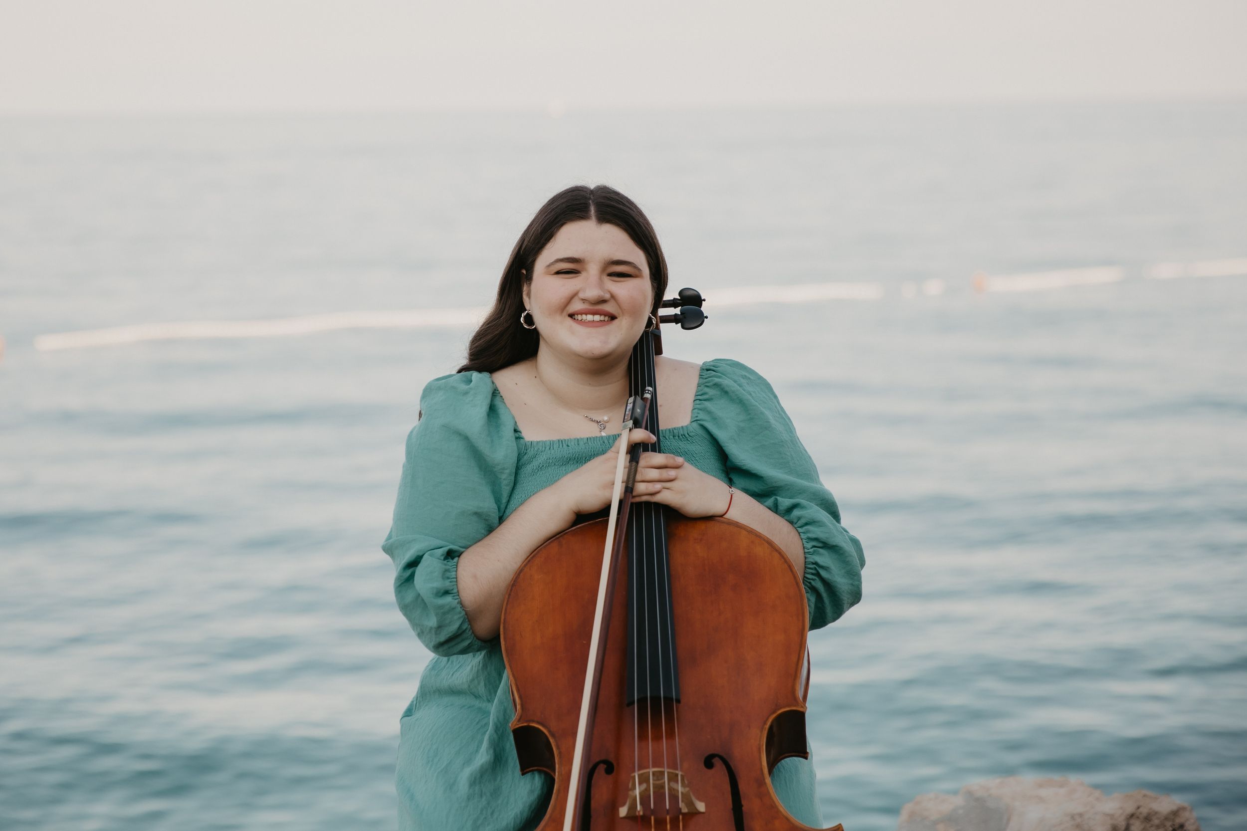 Musician wearing turquoise sweater holds cello against ocean backdrop at sunset.