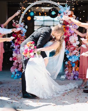 Wedding couple in romantic dip pose under colorful floral arch with confetti falling.