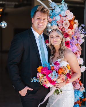 Wedding couple pose in front of vibrant balloon and flower arch backdrop.