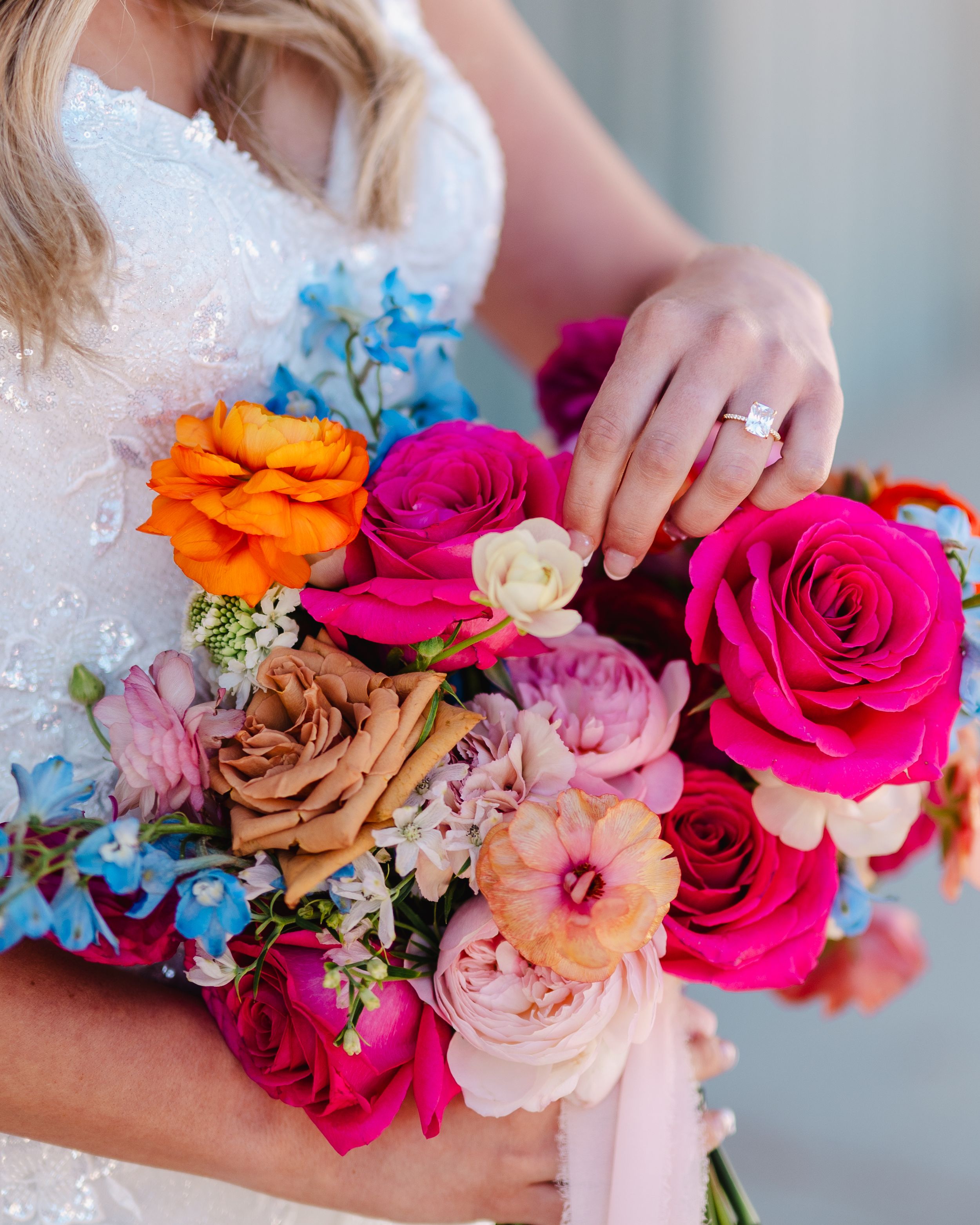 Colorful bridal bouquet with bright roses and blue delphinium held by person in white dress.