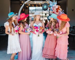 Bridesmaids in pastel dresses and colorful hats laughing together under floral arch.