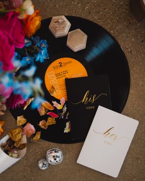Wedding vow books and ring boxes arranged on vinyl record with colorful flower petals.