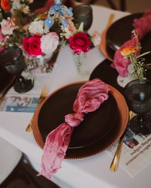 Wedding table setting with pink velvet napkin, dark plates, and vibrant floral centerpieces.