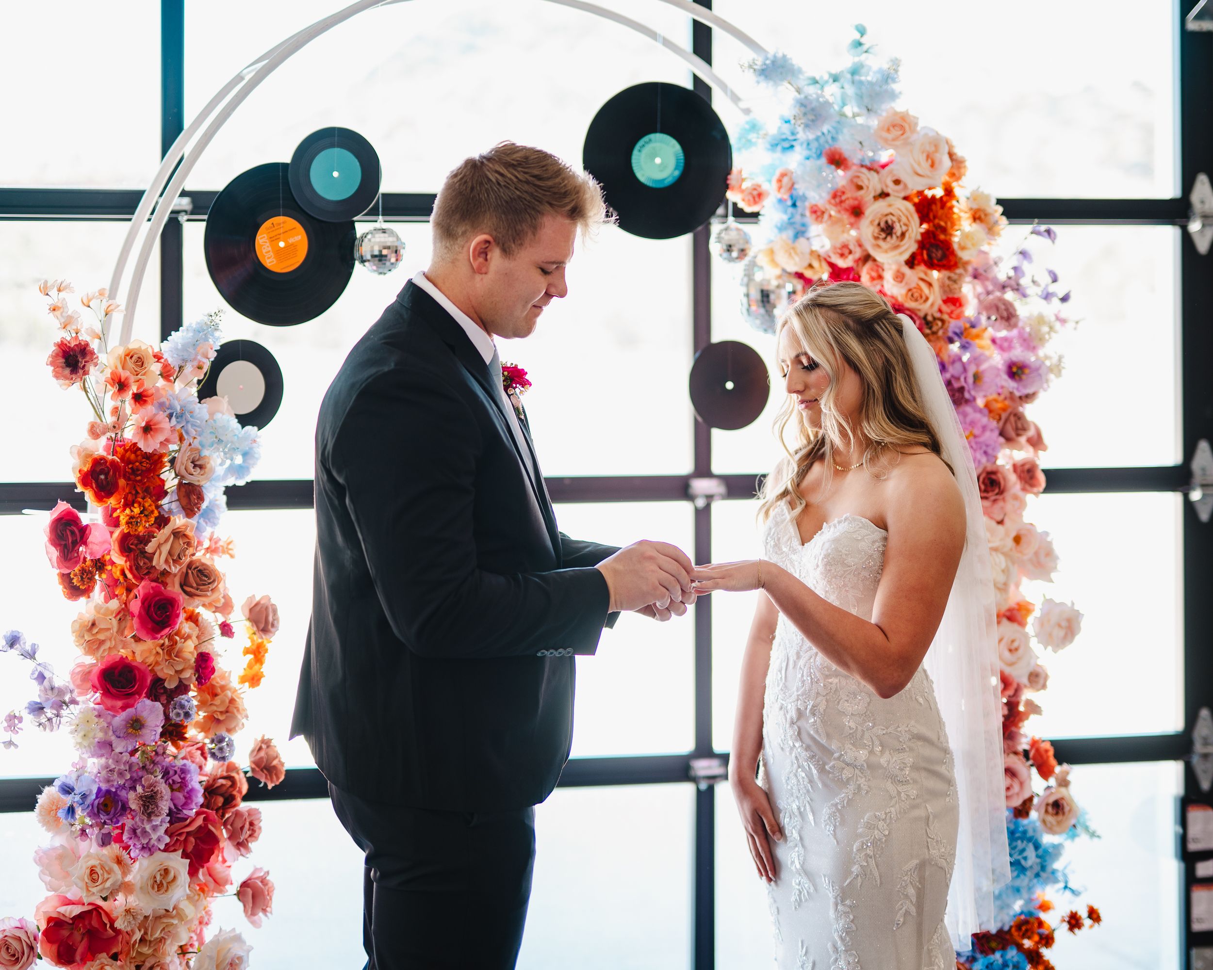 Wedding ceremony with colorful floral arch and vinyl record decor as newlyweds exchange rings.