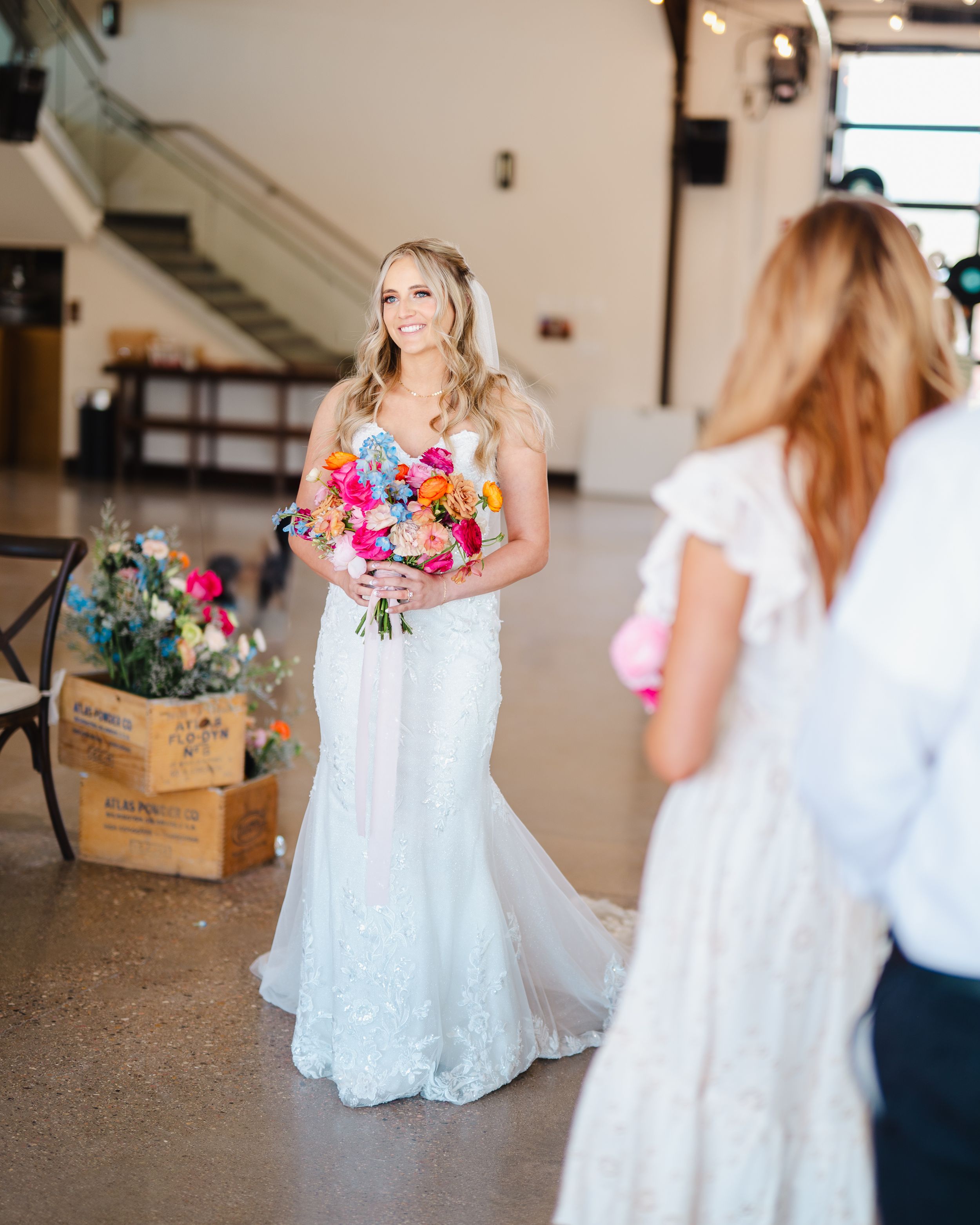 Bride in lace wedding dress holds vibrant multicolored bouquet in industrial venue setting.