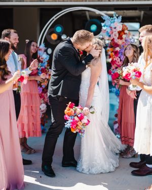Wedding party celebrates as newlyweds share first kiss under arch with pink and blue florals.