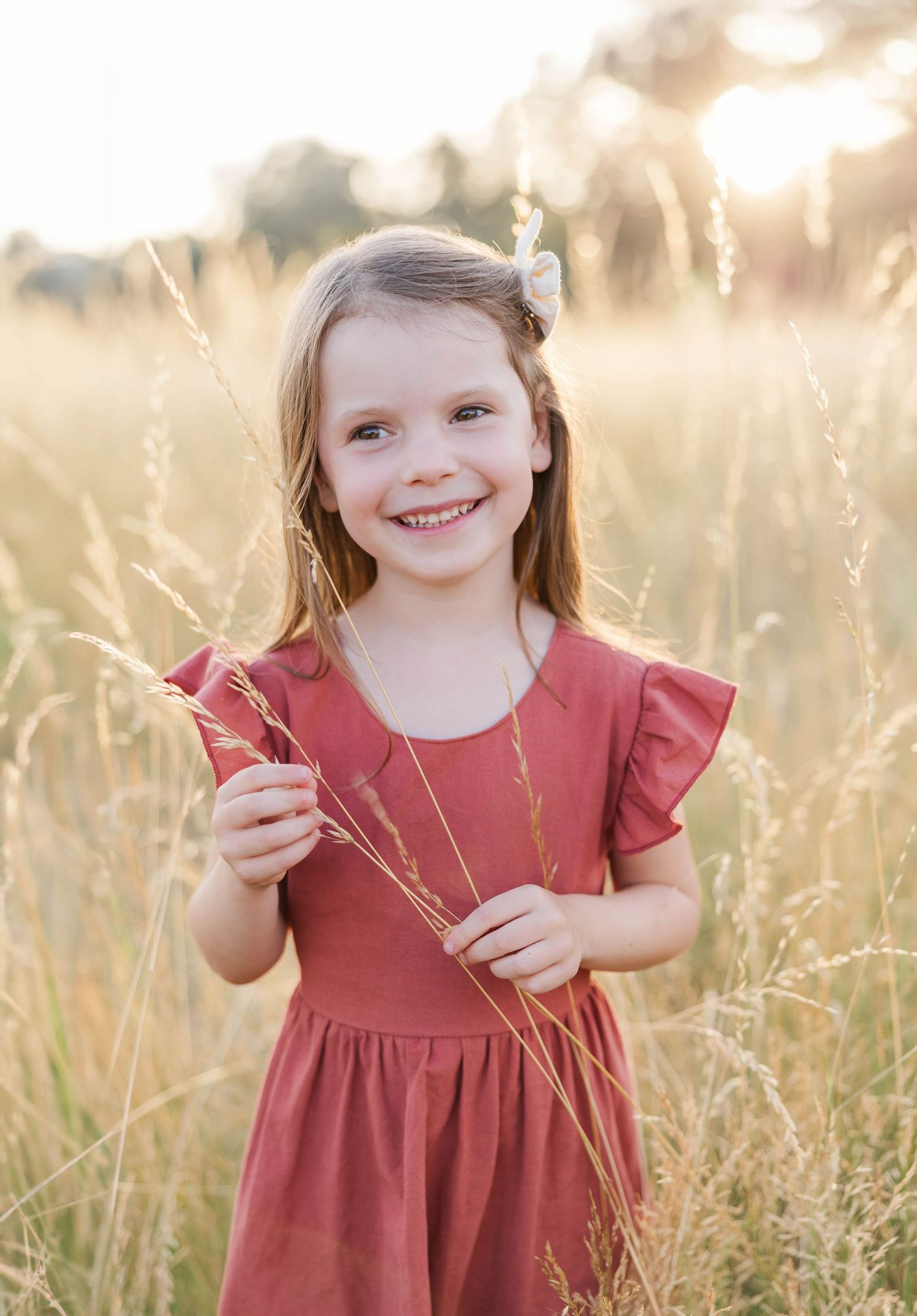 Young girl in red dress smiling in golden field holding tall grass stems in warm sunlight.